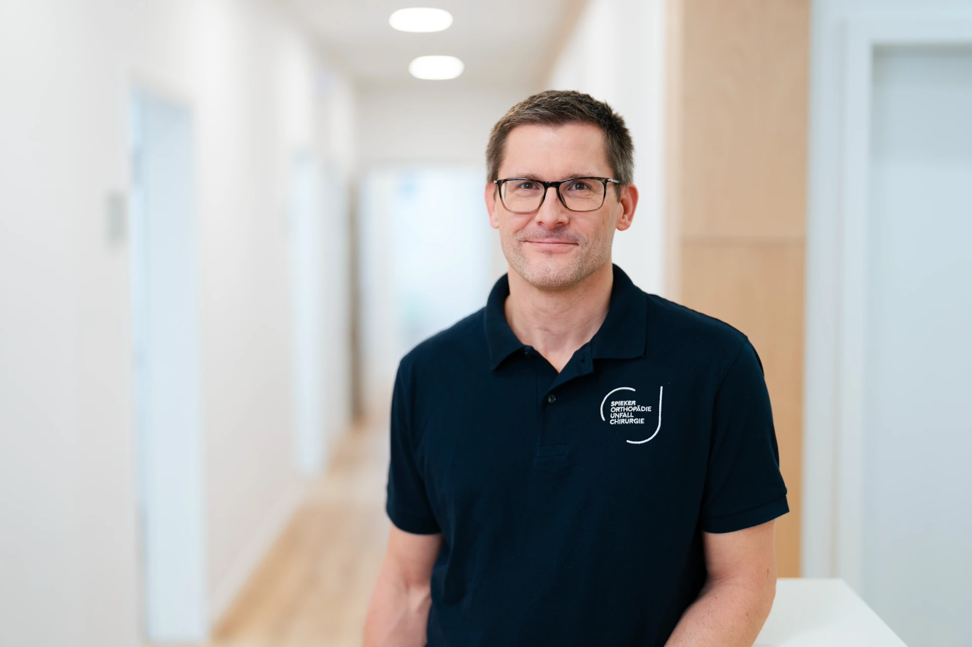 Jan Seden Spieker Mann mit Brille in einem modernen Flur, trägt ein schwarzes Polohemd mit einem Textlogo.