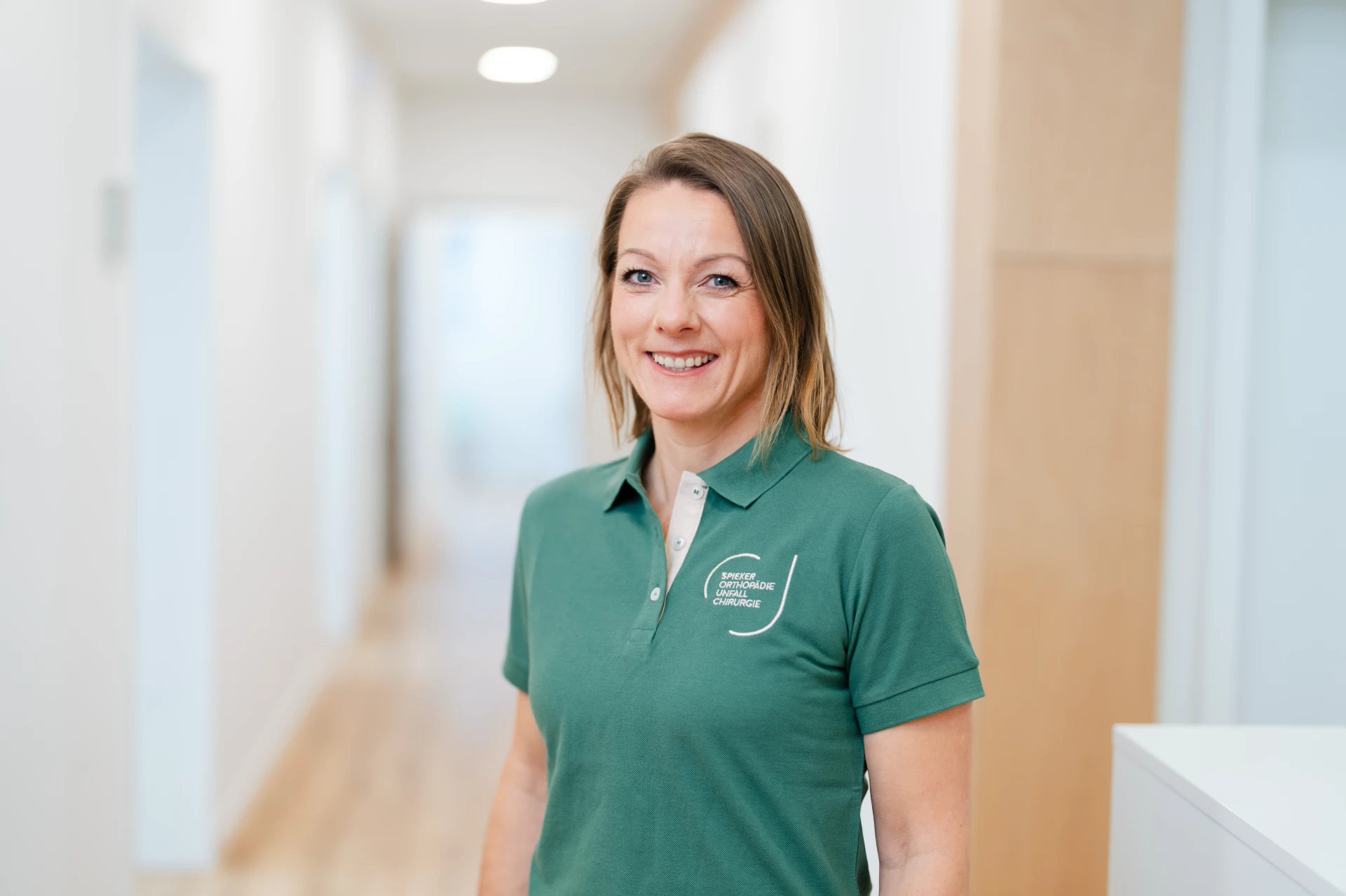 Jessica Nöcker Frau in einem grünen Poloshirt lächelt in einem hellen Flur mit Holzfußboden.