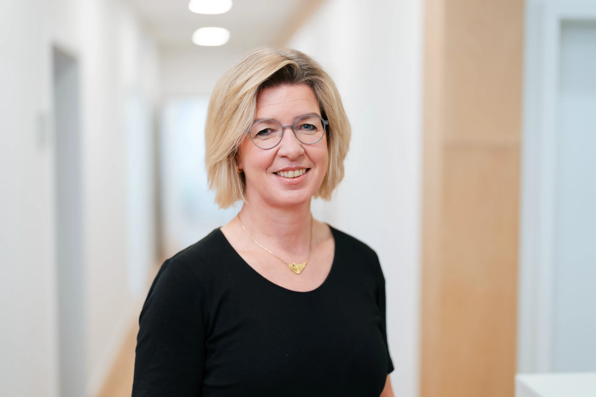 Manuela Ernst Lächelnde Frau mit blonden, schulterlangen Haaren und Brille in einem modernen, hellen Flur.