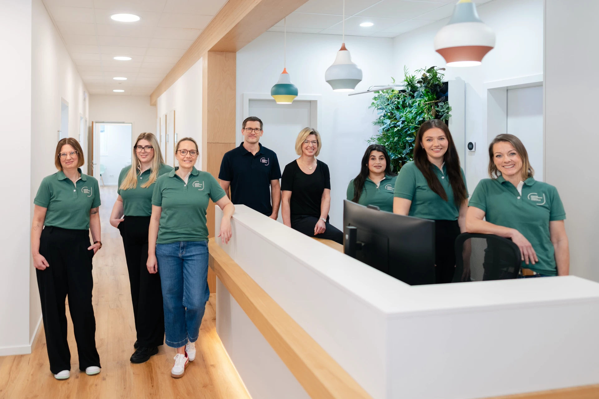 Orthopädie Rietberg | Diagnostik, Therapie & MRT – Spieker Ein Team von sieben Mitarbeitern in grünen Shirts steht hinter einem Empfangstresen in einer modernen Praxis.