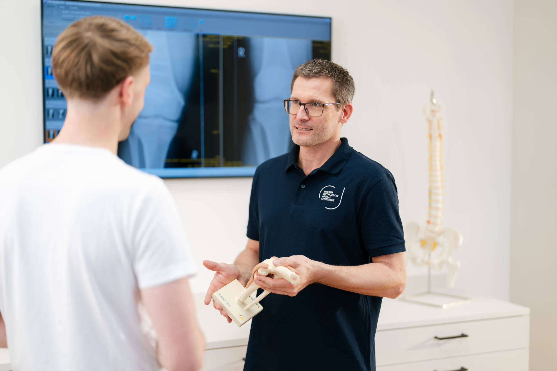 Regenerative Orthopädie | Belastbarkeit zurückgewinnen Ein Fachmann erklärt einem Mann anatomische Modelle von Wirbelsäule und Kniegelenk in einem modernen Raum.