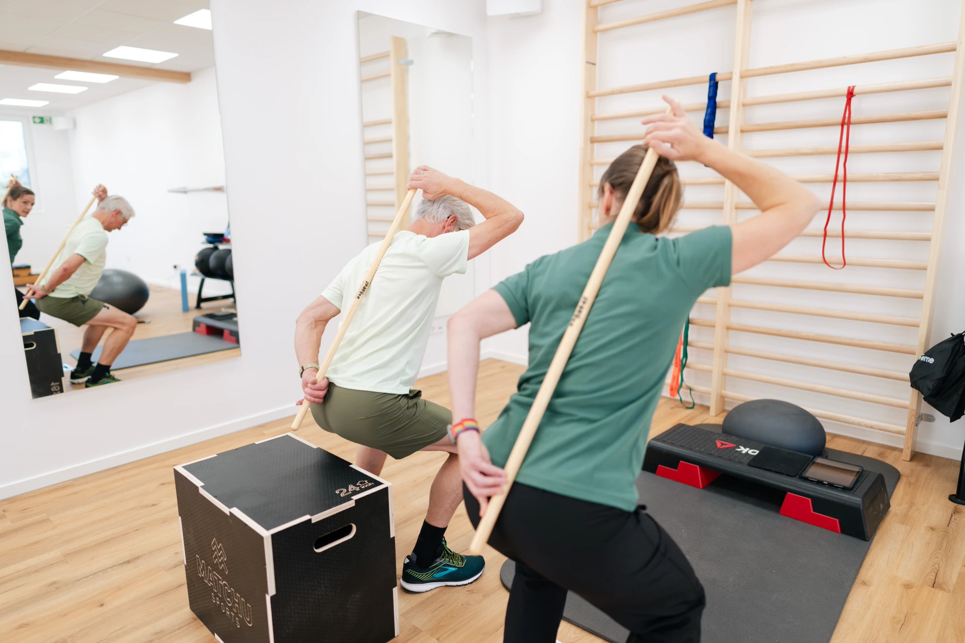 Zwei Personen machen Übungen mit einem Holzstock in einem modernen Trainingsraum.